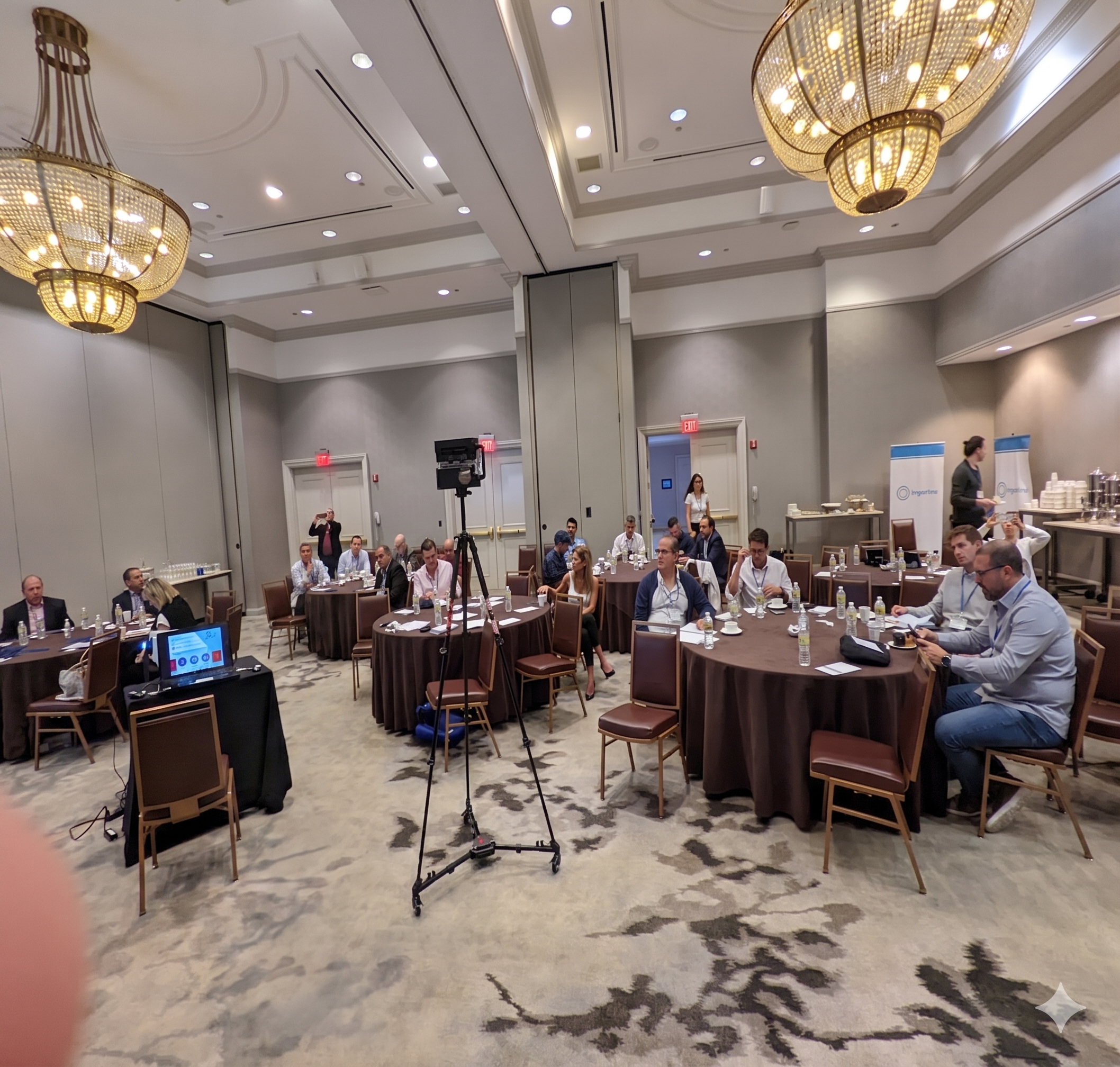 Facilitator at the front of an executive roundtable workshop — participants engaged at tables in a chandelier ballroom